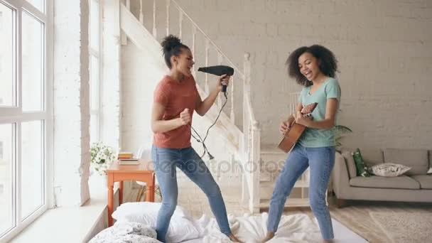 Karışık yarış genç komik kızlar saç kurutma makinesi ile şarkı söylemek ve bir yatakta akustik gitar çalmak dans. Kız kardeş eğleniyor evde yatak odasında eğlence — Stok video