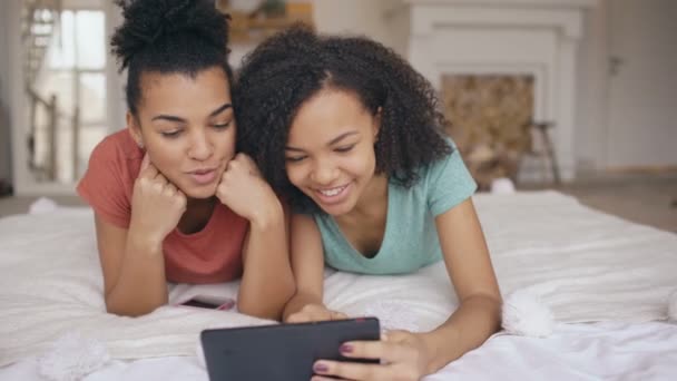 Deux joyeuses petites amies amusantes de race mixte partageant les médias sociaux à l'aide d'un ordinateur tablette et parle couché dans le lit à la maison — Video