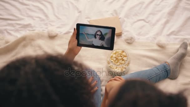 Von oben: zwei Freundinnen sitzen zu Hause auf dem Bett und unterhalten sich per Skype auf einem Tablet-Computer mit ihrem Freund, der Urlaub am Strand macht — Stockvideo