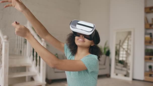 Feliz sorrindo jovem mestiça recebendo experiência usando óculos VR de realidade virtual e mãos em movimento em casa — Vídeo de Stock