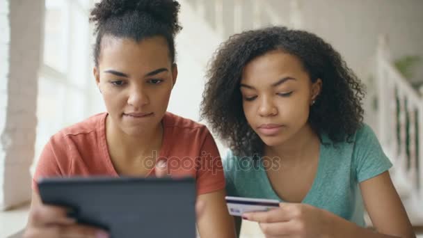 Deux joyeuses copines de course mixte frisée shopping en ligne avec tablette et carte de crédit à la maison — Video