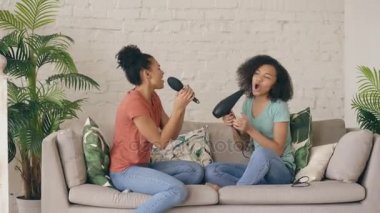 Karışık yarış genç komik kızlar şarkı saç kurutma makinesi ile dans ve koltukta oturan tarak. Kız kardeş eğleniyor eğlence ev concept, oturma odasında