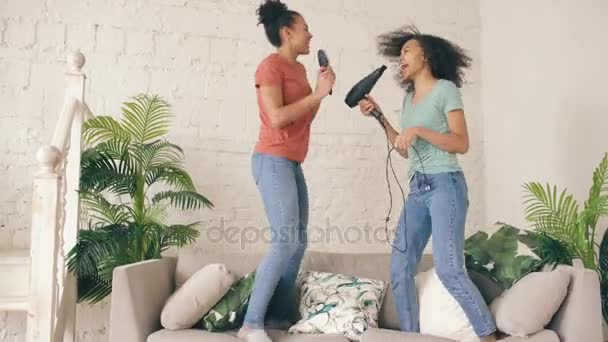 Misto razza giovani ragazze divertenti ballare cantando con asciugacapelli e pettine saltare sul divano. Sorelle che si divertono il tempo libero in salotto a casa concetto — Video Stock