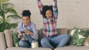 Yavaş hareket iki karma yarış kıvırcık kız kanepede oturan arkadaş gamepad ile konsol bilgisayar oyunları oynamak ve evde eğlenmek