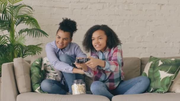 Mouvement lent de deux amis mixtes de fille bouclée de course assis sur le canapé jouer à des jeux informatiques de console avec manette de jeu et amusez-vous à la maison — Video