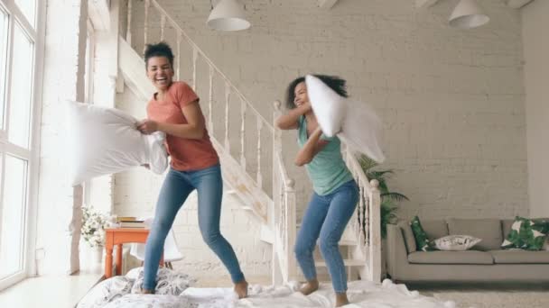 Slow motion van twee gemengd ras jonge mooie meisjes springen op bed en kussens met plezier thuis te bestrijden — Stockvideo