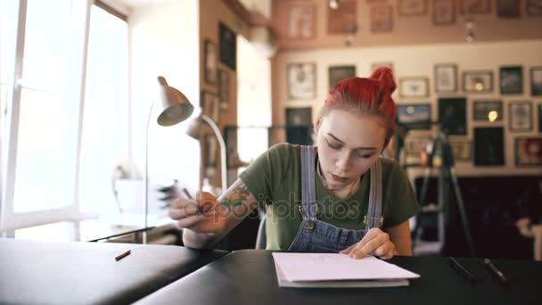 Giovane attraente donna dai capelli rossi tatuatrice seduta a tavola e creare schizzo per il tatuaggio in studio al chiuso — Video Stock
