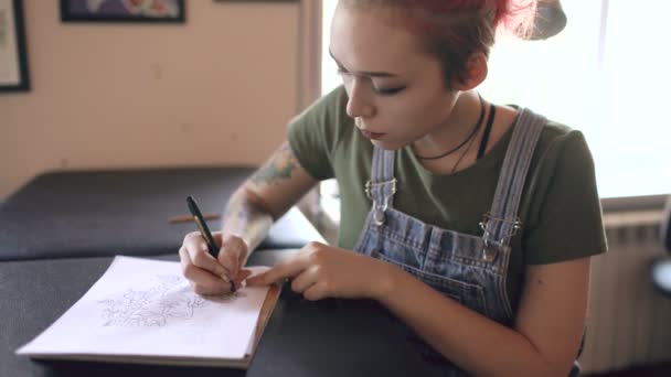 Joven atractiva mujer pelirroja artista del tatuaje sentado en la mesa y la creación de boceto para tatuar en el estudio en interiores — Vídeos de Stock