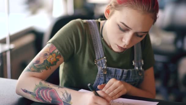 Nahaufnahme einer jungen attraktiven rothaarigen Tätowiererin, die am Tisch sitzt und Skizzen für Tätowierungen im Studio entwirft — Stockvideo