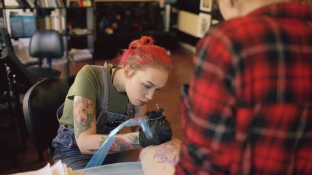 Jovem tatuador de cabelos vermelhos feminino tatuagem imagem na perna do cliente sobre esboço em estúdio dentro de casa — Vídeo de Stock