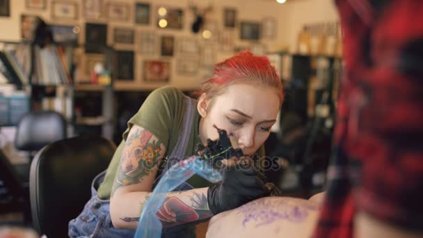 Joven mujer pelirroja tatuaje artista tatuaje imagen en la pierna del cliente sobre boceto en el estudio en interiores — Vídeo de stock