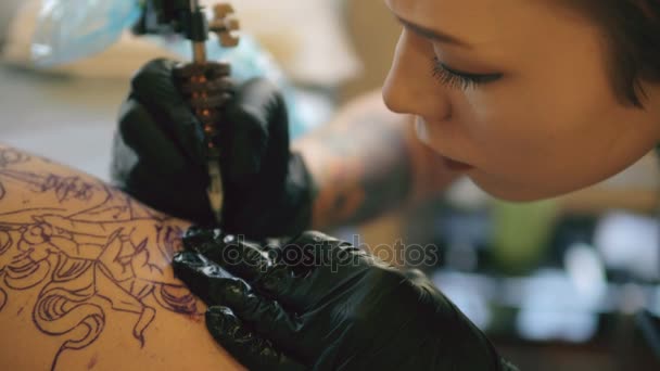 Closeup μικρά κόκκινα μαλλιά τατουάζ γυναίκα καλλιτέχνης τατουάζ εικόνα στο πόδι του πελάτη πάνω από σκίτσο στο studio σε εσωτερικούς χώρους — Αρχείο Βίντεο