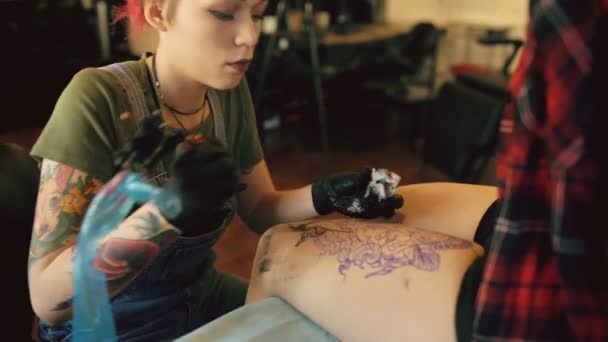 Red haired girl tattoo artist tattooing picture on lef of young girl client in studio — Stock Video