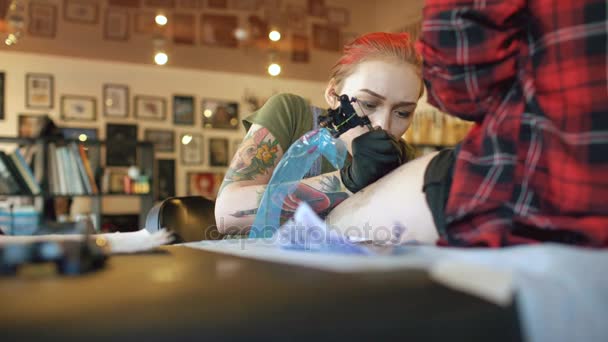 Tilt up disparar de joven pelirroja mujer tatuaje artista tatuaje imagen en la pierna de joven chica cliente en el estudio — Vídeos de Stock