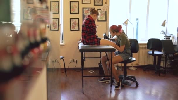 Unga kvinnliga röda haired tatuerare tatuera bild på ben av klienten över skiss i studio inomhus — Stockvideo