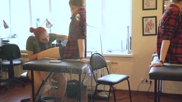 Gyönyörű vörös hajú nő tetováló művész tetoválás kép, a lábát a fiatal lány ügyfél stúdióban — Stock videók