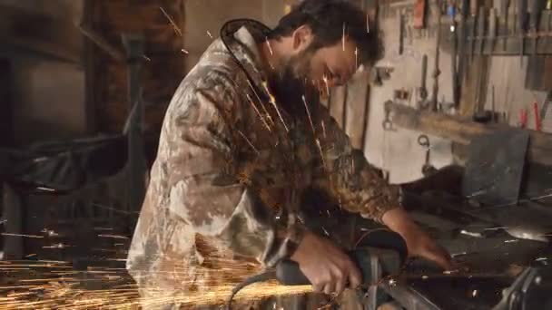Jeune homme barbu utilisant meuleuse d'angle pour travailler sur un morceau de métal dans un atelier de forge, étincelles de broyage volant autour — Video