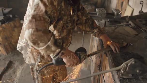 Jovem barbudo usando moedor de ângulo para trabalhar em peça de metal em oficina de ferreiro, moagem faíscas voando ao redor — Vídeo de Stock