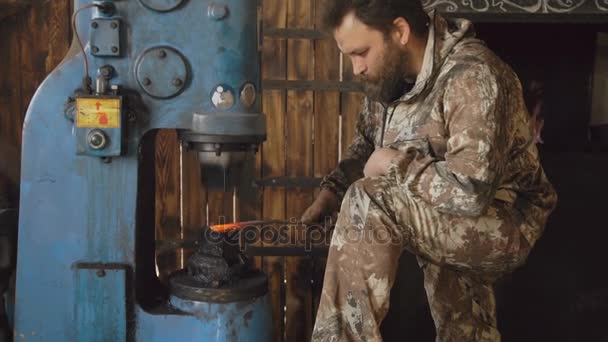 Γενειοφόρος νεαρός σιδεράς χρησιμοποιώντας υδραυλικό πιεστήριο για την παραγωγή χάλυβα όπλων στο εργαστήρι smithy — Αρχείο Βίντεο