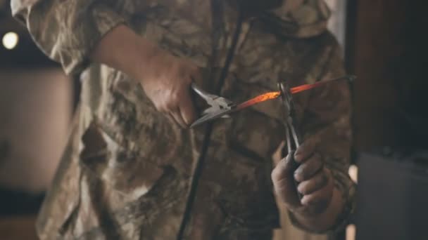 Крупный план кузнечных рук, сгибающих горячий металлический нож плоскогубцами в традиционном кузнице — стоковое видео