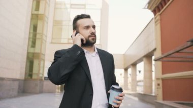 Genç iş adamı modern ofis binaları yakınındaki kahve Kupası ile anlaşmalar yapmaya cep telefonuyla konuşan sakallı