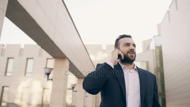 Dolly tiro de homem de negócios falando sobre telefone celular fazendo negócios e caminhando perto de edifícios de escritórios modernos — Vídeo de Stock