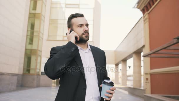Jovem homem de negócios barbudo falando no telefone celular fazendo negócios com xícara de café perto de edifícios de escritórios modernos — Vídeo de Stock