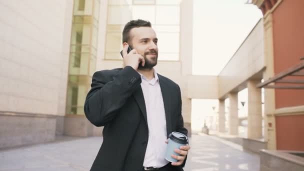 Dolly disparo de un joven hombre de negocios barbudo hablando en el teléfono inteligente con una taza de café cerca de modernos edificios de oficinas — Vídeos de Stock