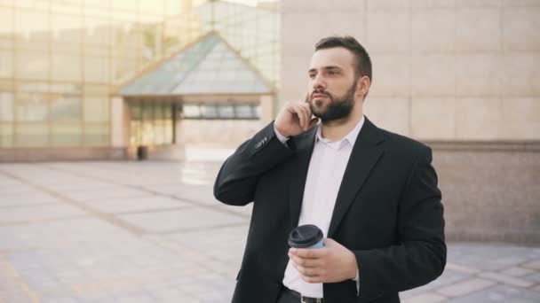 Young bearded business man talking at smartphone with cup of coffee near modern office buildings — Stock Video