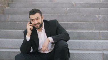 Genç iş adamı sokakta merdivenlerde oturan anlaşmalar yapmaya cep telefonuyla konuşan sakallı