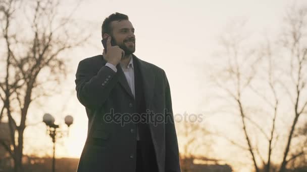 Joven hombre de negocios barbudo con abrigo hablando en el teléfono inteligente y haciendo ofertas en la calle con luz solar retroiluminada en el tiempo de la noche — Vídeos de Stock