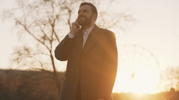 Молодой бородатый бизнесмен в пальто разговаривает на смартфоне и заключает сделки на улице с подсветкой солнечного света в вечернее время — стоковое видео