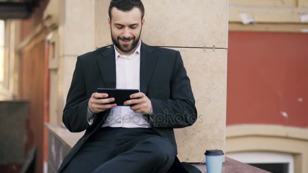 Молодой бородатый бизнесмен читает новости на планшетном компьютере и пьет кофе во время перерыва возле своего офисного здания — стоковое видео