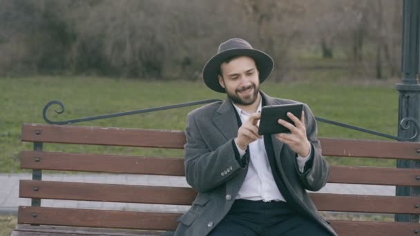 Hipser creatief zakenman in hoed met behulp van tablet pc-computer en zittend op stad straat bankje in park tijdens pauze — Stockvideo