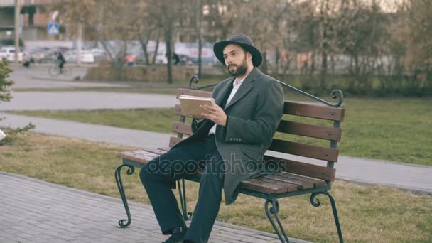 Seorang seniman kreatif Hipser dengan topi dan mantel menggambar dengan pena dan buku sketsa duduk di bangku jalan kota di taman — Stok Video