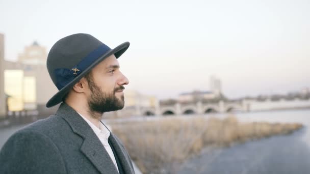 Joven hombre barbudo turista en sombrero y abrigo mirando paisaje urbano y soñando despierto mientras está parado a orillas del río — Vídeos de Stock