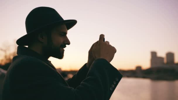 夕暮れ時彼のスマート フォンのカメラで街のスカイラインのパノラマ写真を撮影の帽子の旅行者の男のシルエット — ストック動画