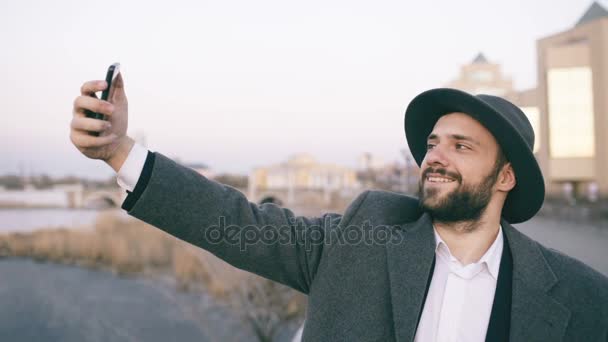 Giovane uomo turistico felice in ahat e cappotto sorridente mentre scattare foto selfie con il telefono cellulare sulla riva del fiume della città — Video Stock