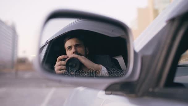 Reflexión en el espejo lateral del joven detective privado sentado dentro del coche y fotografiando con la cámara dslr — Vídeos de Stock