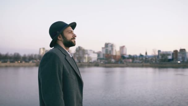 Молодой бородатый турист в шляпе и пальто смотрит городской пейзаж и мечтает, стоя на берегу реки — стоковое видео