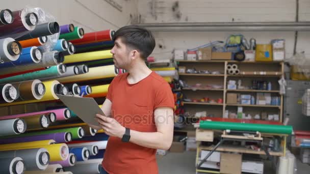 Dolly disparo de trabajador joven en el almacén con el inventario de comprobación portapapeles. El hombre trabaja en el departamento de ventas de materiales publicitarios — Vídeo de stock