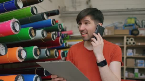 Dolly colpo di lavoratore sorridente in magazzino parlando smartphone e controllando l'inventario. Uomo lavora nel reparto vendite di materiale pubblicitario — Video Stock