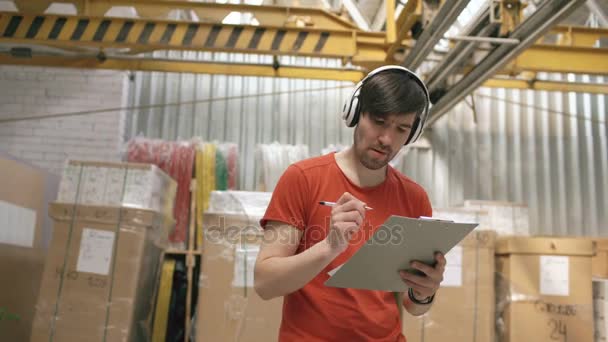 Dolly záběr šťastný mladý dělník v průmyslových skladu, poslouchá hudbu a tanec během práce. Muž v sluchátka se bavit na pracovišti. — Stock video