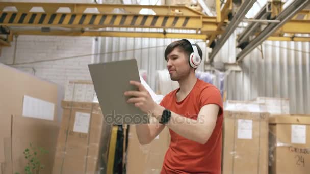 Dolly scatto di Happy giovane operaio in magazzino industriale ascoltando musica e ballando durante il lavoro. Uomo in cuffia divertirsi sul posto di lavoro . — Video Stock