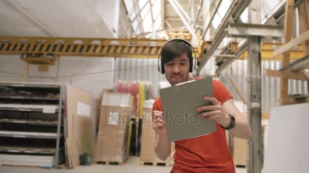 Dolly rodó de Happy joven trabajador en almacén industrial escuchando música y bailando durante el trabajo. Hombre en auriculares divertirse en el lugar de trabajo . — Vídeos de Stock