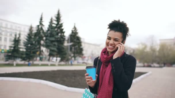 Dolly shot Attrayant mixte femme d'affaires parlant smartphone et boire du café promenades dans la rue de la ville avec des sacs à provisions — Video