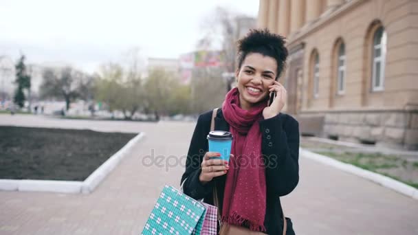 Dolly shot of Attractive mixed race business woman talking smartphone and drinking coffee walks in city street with shopping bags — Stock Video