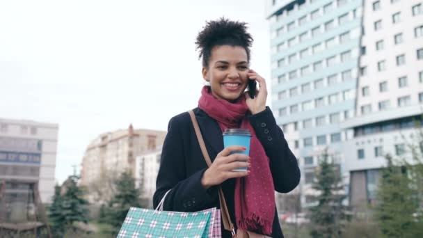 Atractiva mujer de negocios de raza mixta hablando teléfono inteligente y beber café en la calle de la ciudad con bolsas de compras — Vídeos de Stock