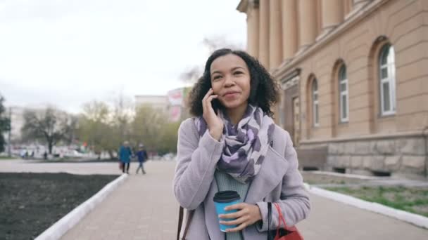 Κουκλίτσα βολή από ελκυστικό μικτή φυλή κορίτσι μιλάμε smartphone και πίνοντας καφέ περπατάει στο δρόμο με τις τσάντες της πόλης. Νεαρή γυναίκα περπάτημα μετά τις αγορές σας στο εμπορικό κέντρο πώλησης — Αρχείο Βίντεο