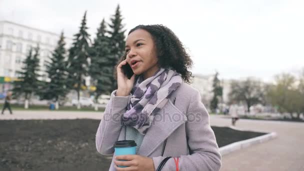 Dolly tiro de menina de raça mista atraente falando smartphone e beber café anda na rua da cidade com sacos. Jovem mulher andando depois de fazer compras na venda do shopping — Vídeo de Stock